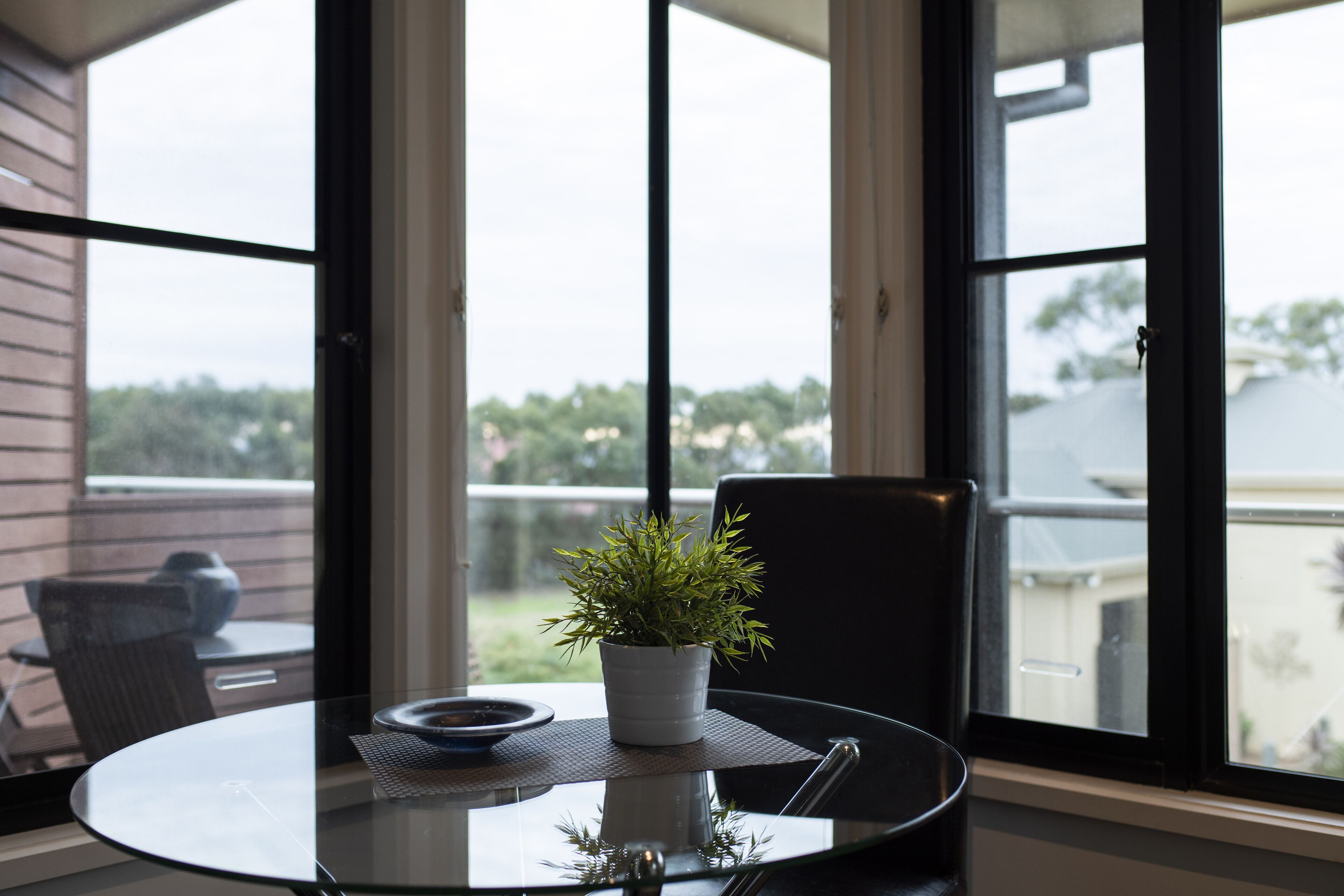 A glass table with a potted plant, a bowl, and a chair in a room with large windows.