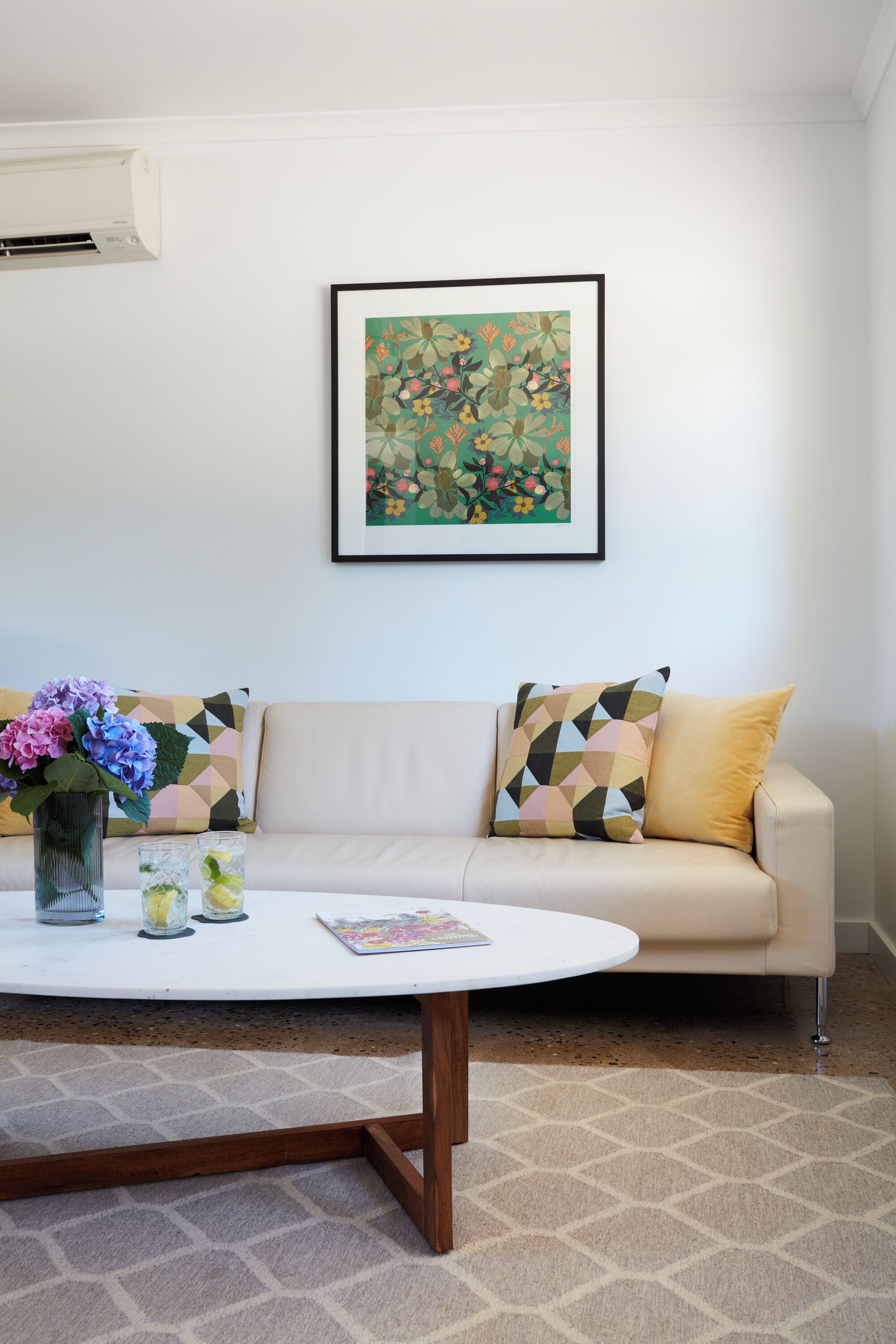 Un salón con un sofá color beige, una mesa redonda de centro, una obra de arte floral enmarcada y un jarrón con flores.