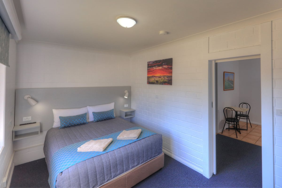 A bedroom with a bed, bedside tables, a nightstand, a painting, and a chair in a hallway.