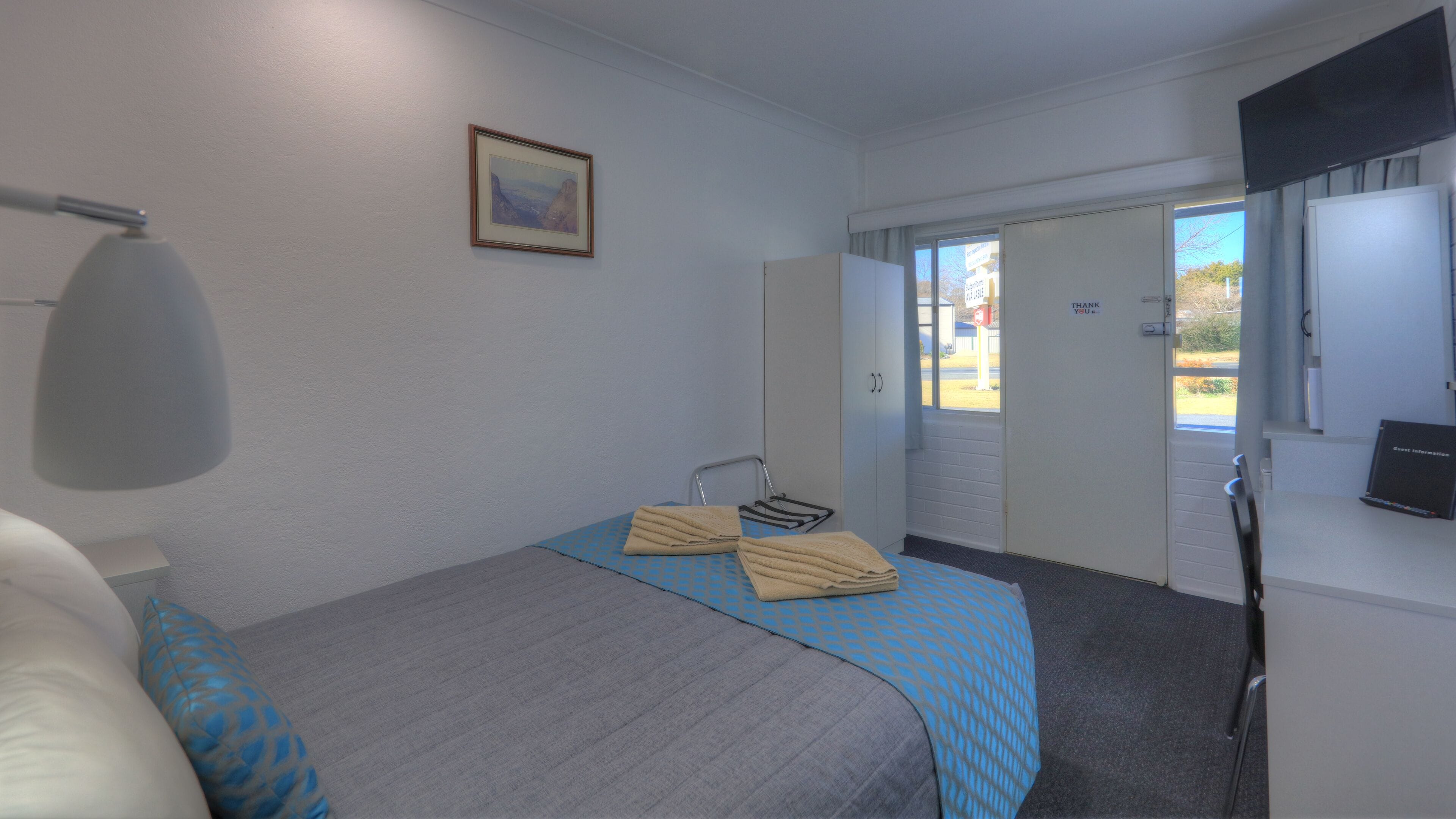 A hotel room with a bed, a desk, a chair, a lamp, a framed picture on the wall, and a window with a view of the outside.