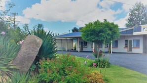 A motel with a covered parking area, a landscaped garden with various plants and flowers, and a clear blue sky with clouds. - Glen Innes Motel (Glen Innes)