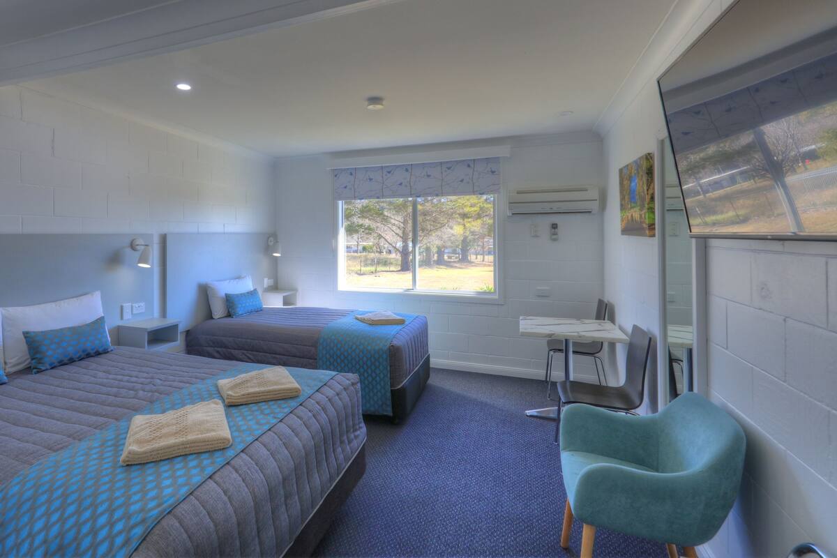 A hotel room with two beds, a desk, a chair, and a large window offering a view of the outdoors.