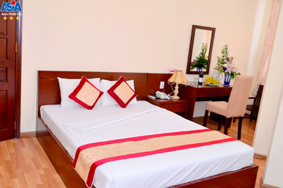 A hotel room with a bed, a desk, a chair, a lamp, and a vase with flowers.