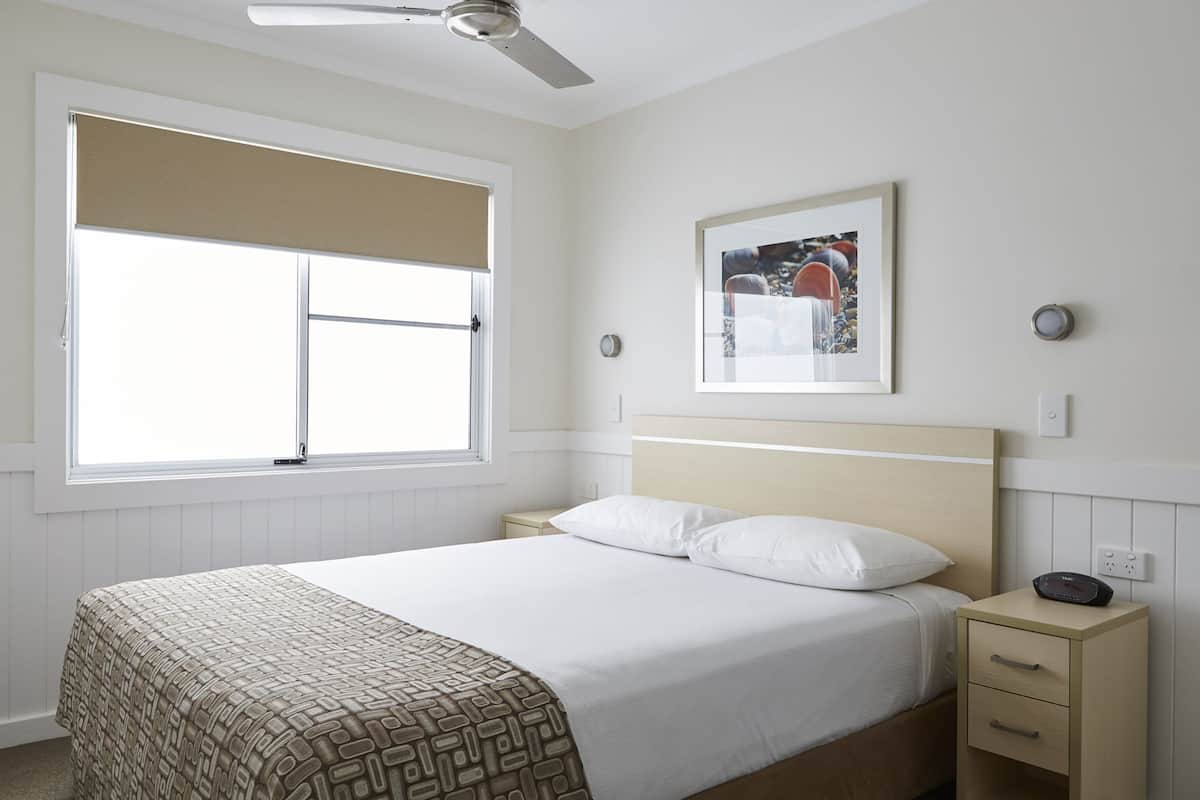 Une chambre à coucher comprenant un lit, une table de chevet, une commode, un ventilateur de plafond, une fenêtre avec des stores et un tableau encadré accroché au mur.