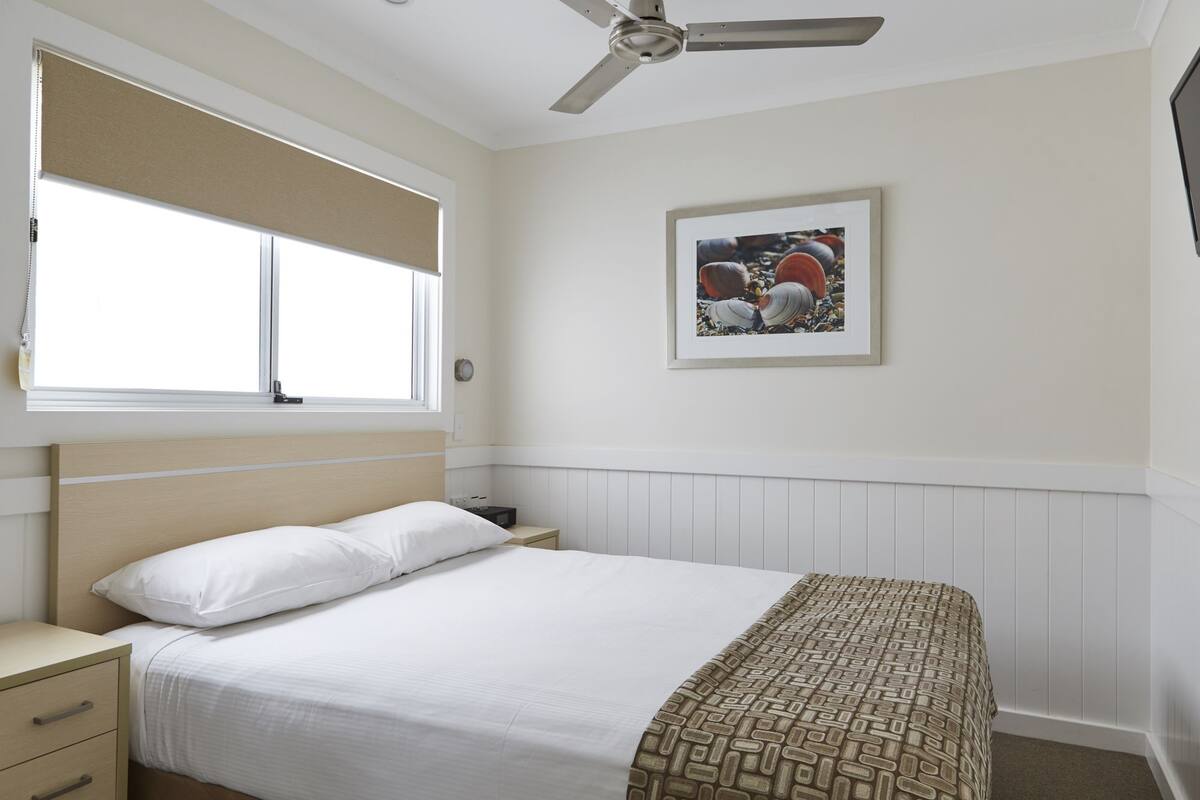 Une chambre à coucher comprenant un lit, une table de chevet, une fenêtre avec des stores, un ventilateur de plafond et un tableau encadré accroché au mur.
