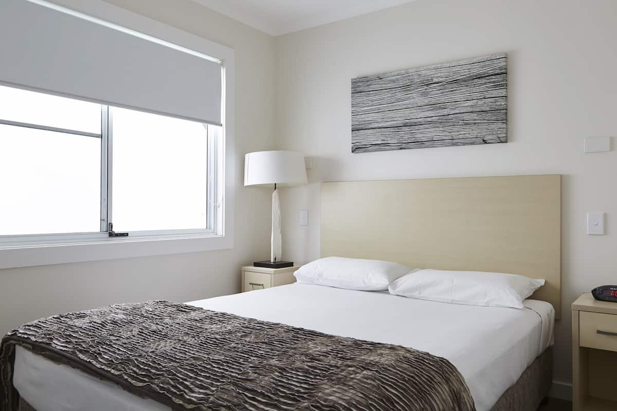 Une chambre d’hôtel comprenant un lit, une table de chevet, une lampe, une fenêtre et une œuvre d’art accrochée au mur.