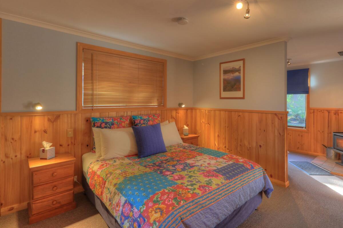 A bedroom with a wooden headboard, a bed with a colorful quilt, a nightstand with a tissue box, a wooden dresser, a window with blinds, a painting on the wall, and a wood-burning stove in the corner.