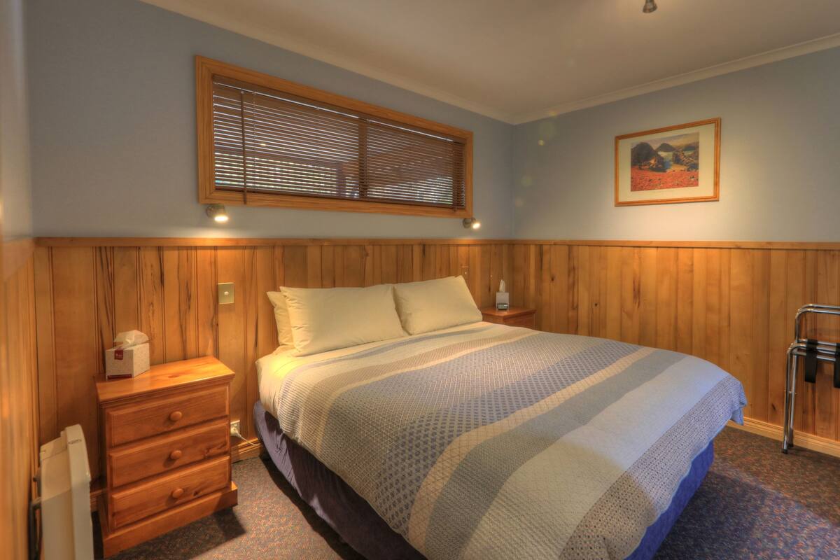A bedroom with a bed, wooden headboard, a nightstand, a drawer unit, a window with blinds, a picture on the wall, and a radiator.