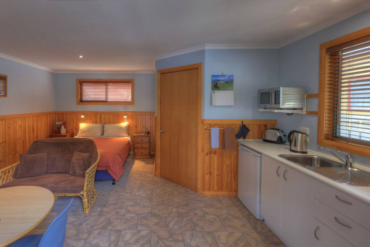 A compact room with a bed, a small dining table, a kitchenette, and a wooden chair.