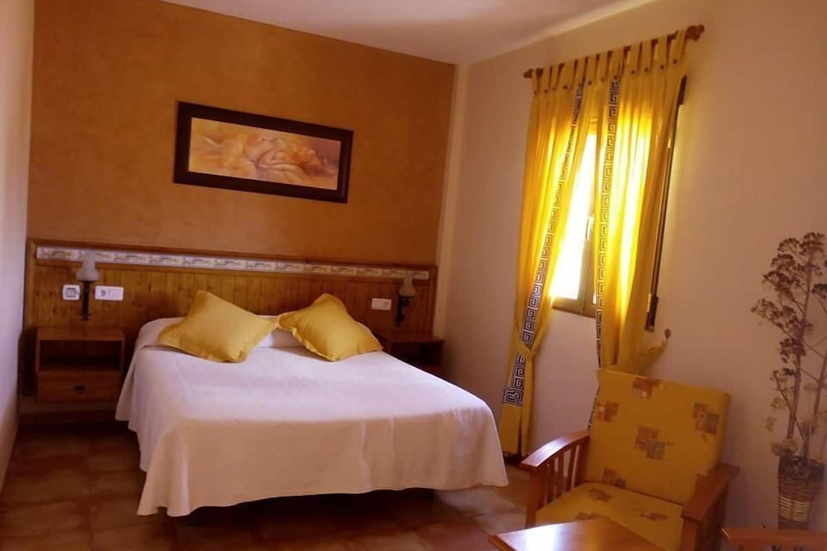 A hotel room with a bed, a chair, a table, and a painting on the wall.