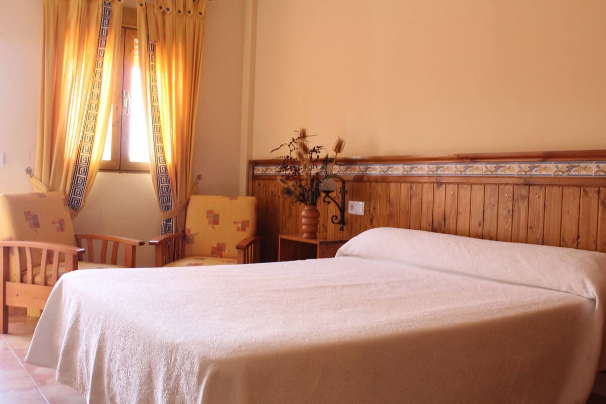 A hotel room with a large bed, wooden paneling, a small table with a vase, and two chairs.