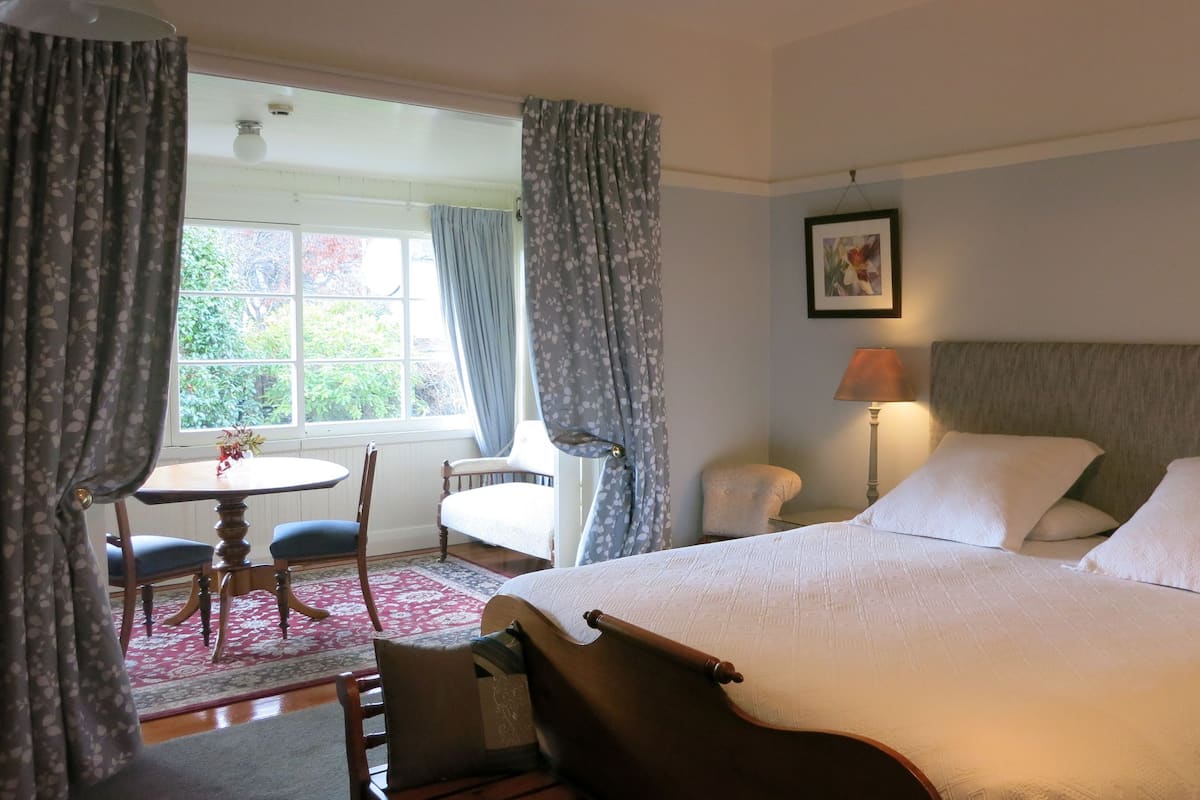 Quarto de hotel com uma cama grande, uma mesa redonda pequena, duas cadeiras, um abajur e uma janela com cortinas.