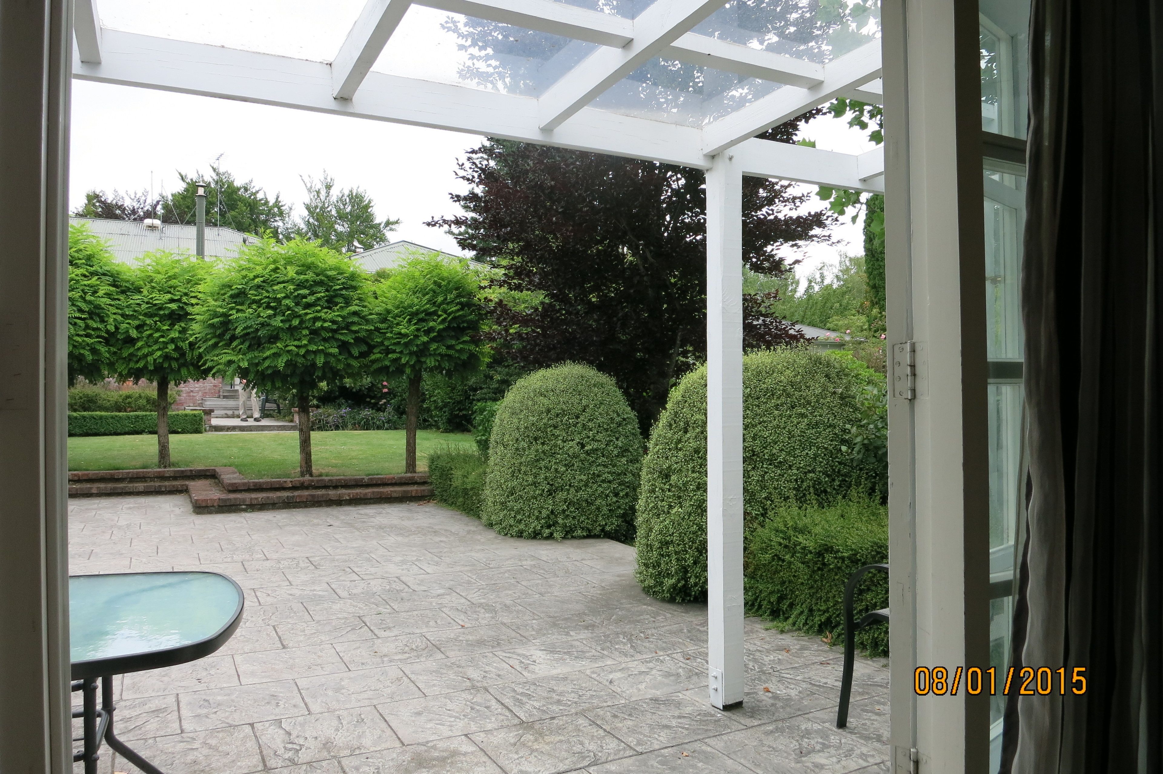 A covered patio area with a stone floor, surrounded by neatly trimmed hedges and trees.