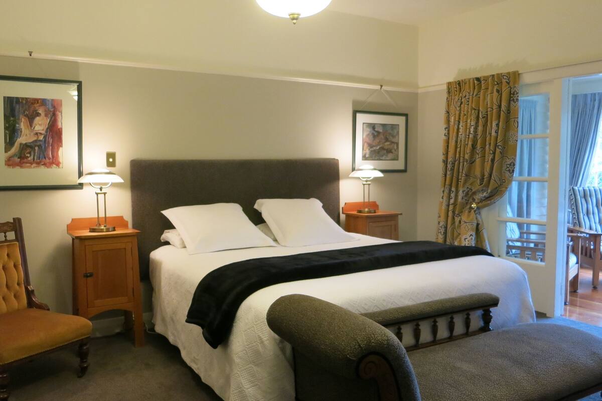 Quarto de hotel com uma cama grande, duas mesas de cabeceira com luminárias, uma cadeira, um banco e duas imagens emolduradas nas paredes.