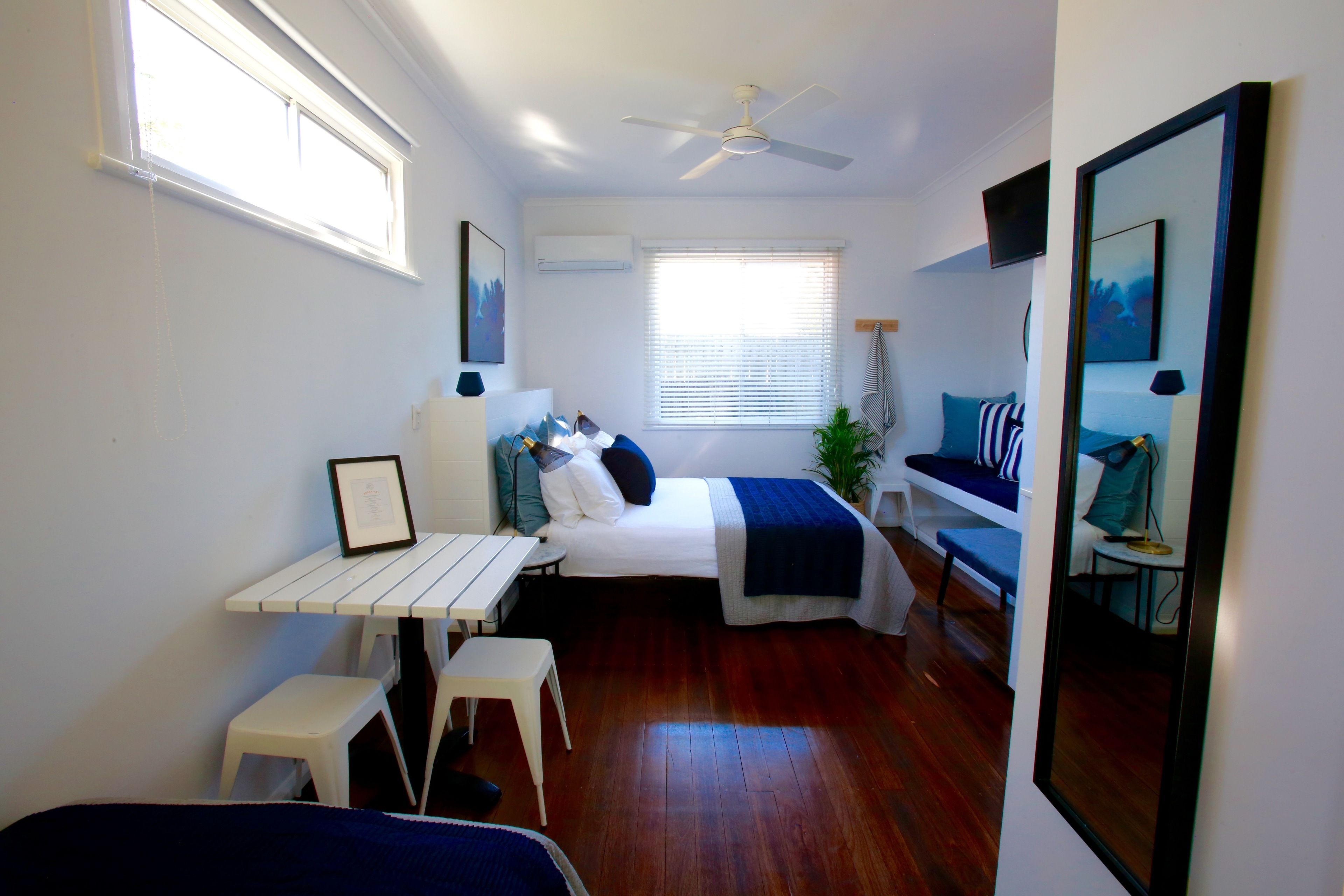 Un dormitorio compacto con cama, mesita de noche y un escritorio pequeño con silla. Hay una ventana con persianas, un ventilador de techo y un espejo en la pared.