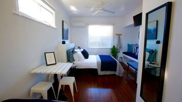 Un dormitorio compacto con cama, mesita de noche y un escritorio pequeño con silla. Hay una ventana con persianas, un ventilador de techo y un espejo en la pared.
