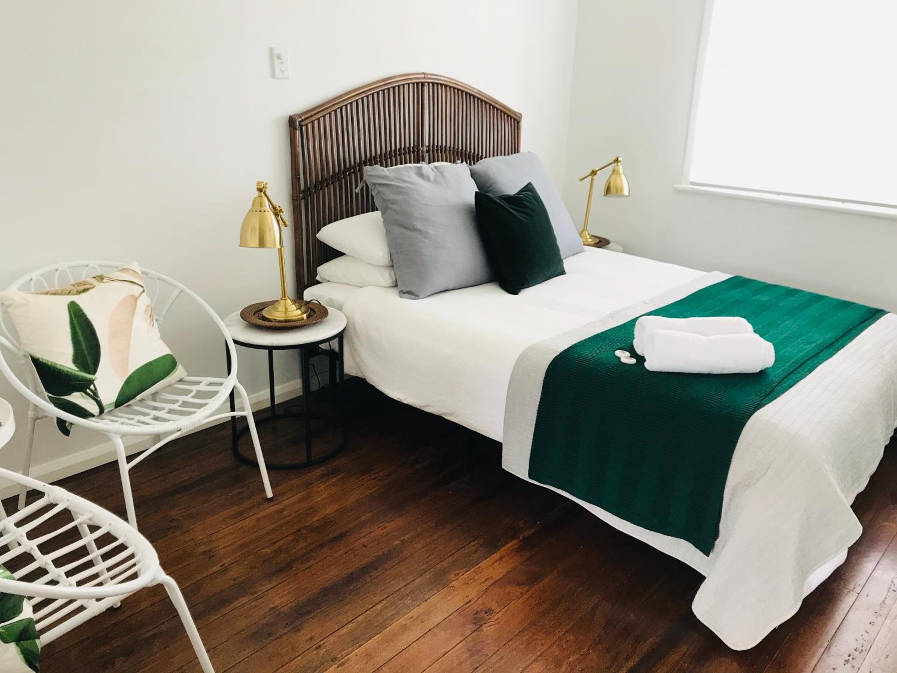 A neatly arranged bedroom with a wooden headboard, a white bedspread, a green throw blanket, and a white chair with a plant.