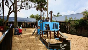 Un parque infantil con una estructura de juego tipo barco pirata, rodeado de arena y árboles.