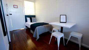 Een kleine, schone kamer met een bed, een bureau en twee witte krukken.