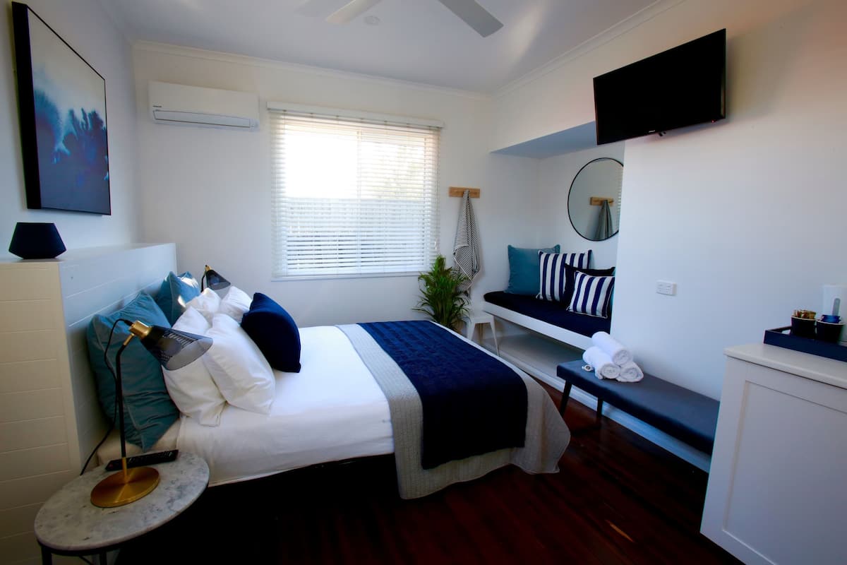 A bedroom with a bed, bedside table, TV, and a window with blinds.