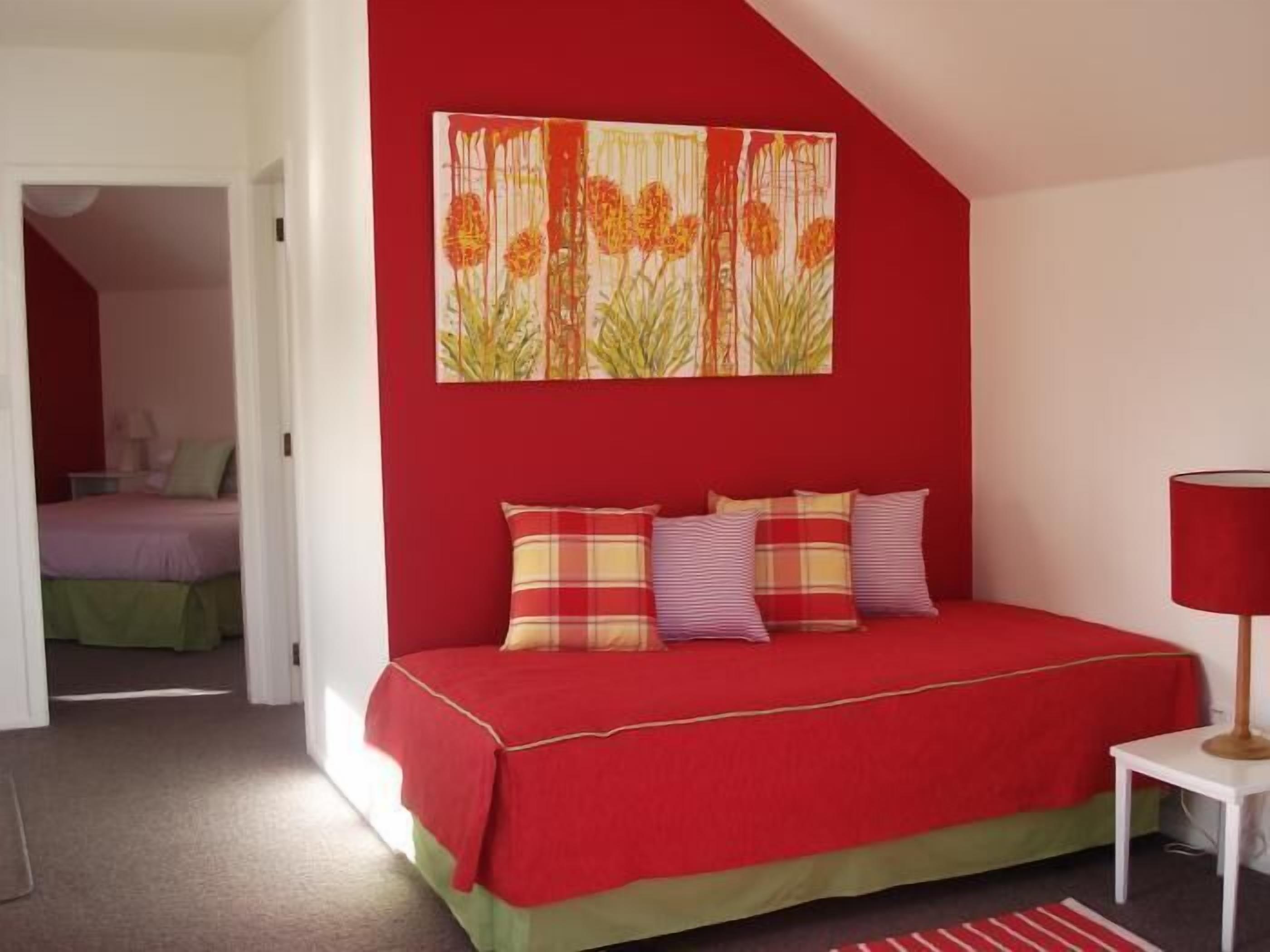 Een slaapkamer met een rode muur, een bed met een sprei in rood en groen, en een schilderij van bloemen.