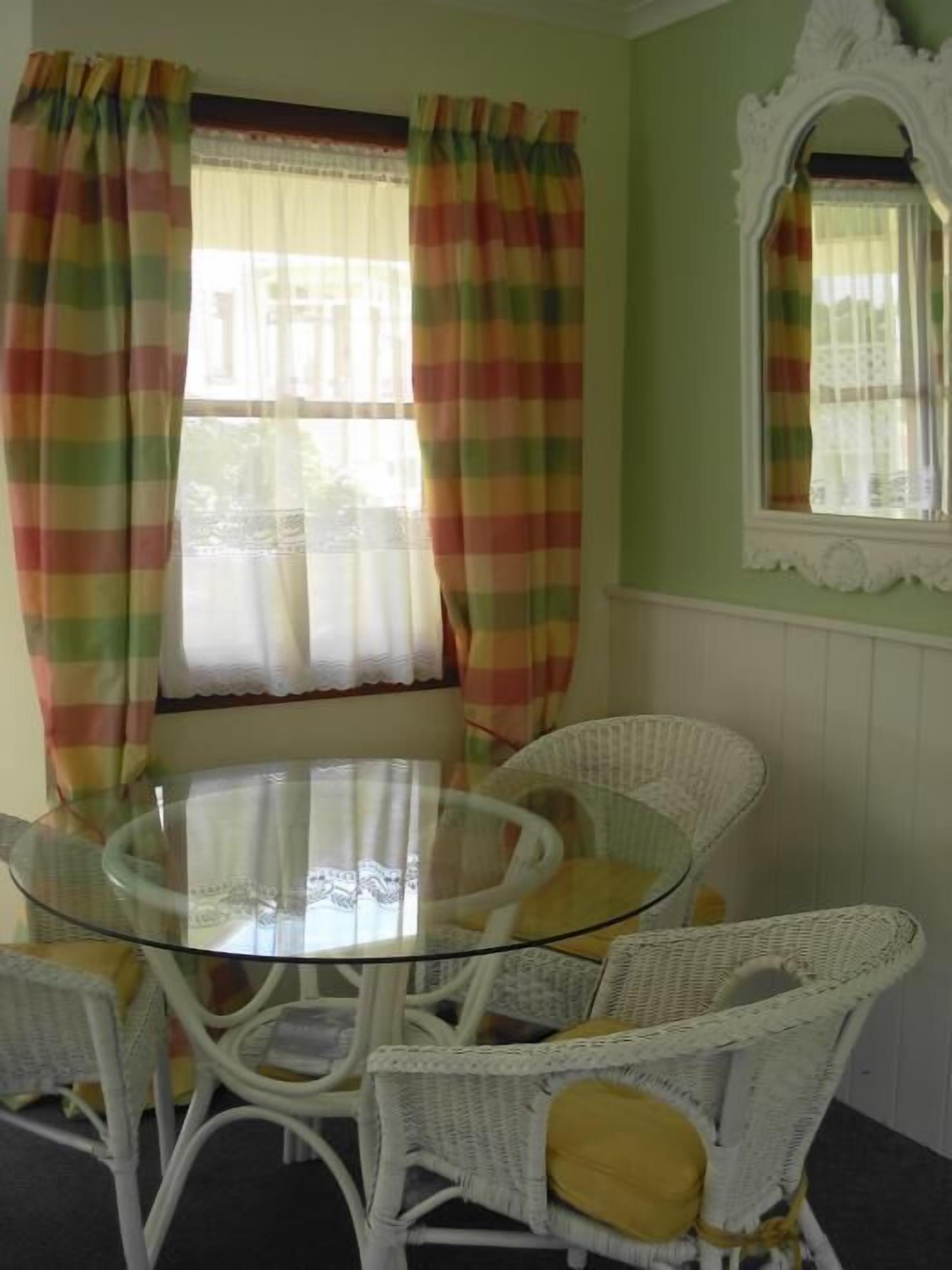 A round glass table with white wicker chairs, a window with plaid curtains, and a decorative mirror on the wall.