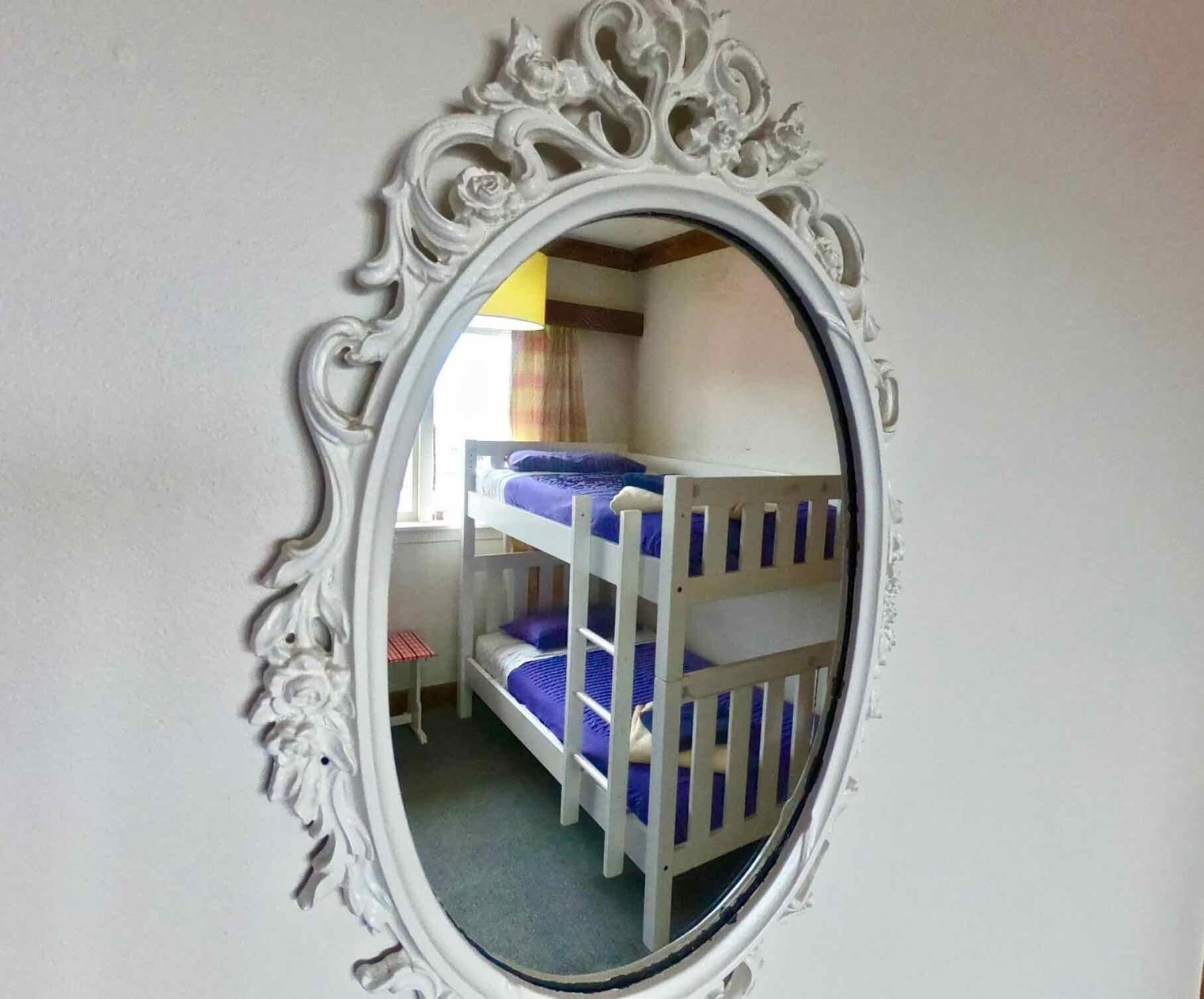 A bunk bed with a white frame and blue bedding, a small desk with a chair, and a mirror with an ornate frame.