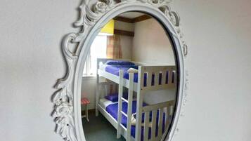 A bunk bed with a white frame and blue bedding, a small desk with a chair, and a mirror with an ornate frame.