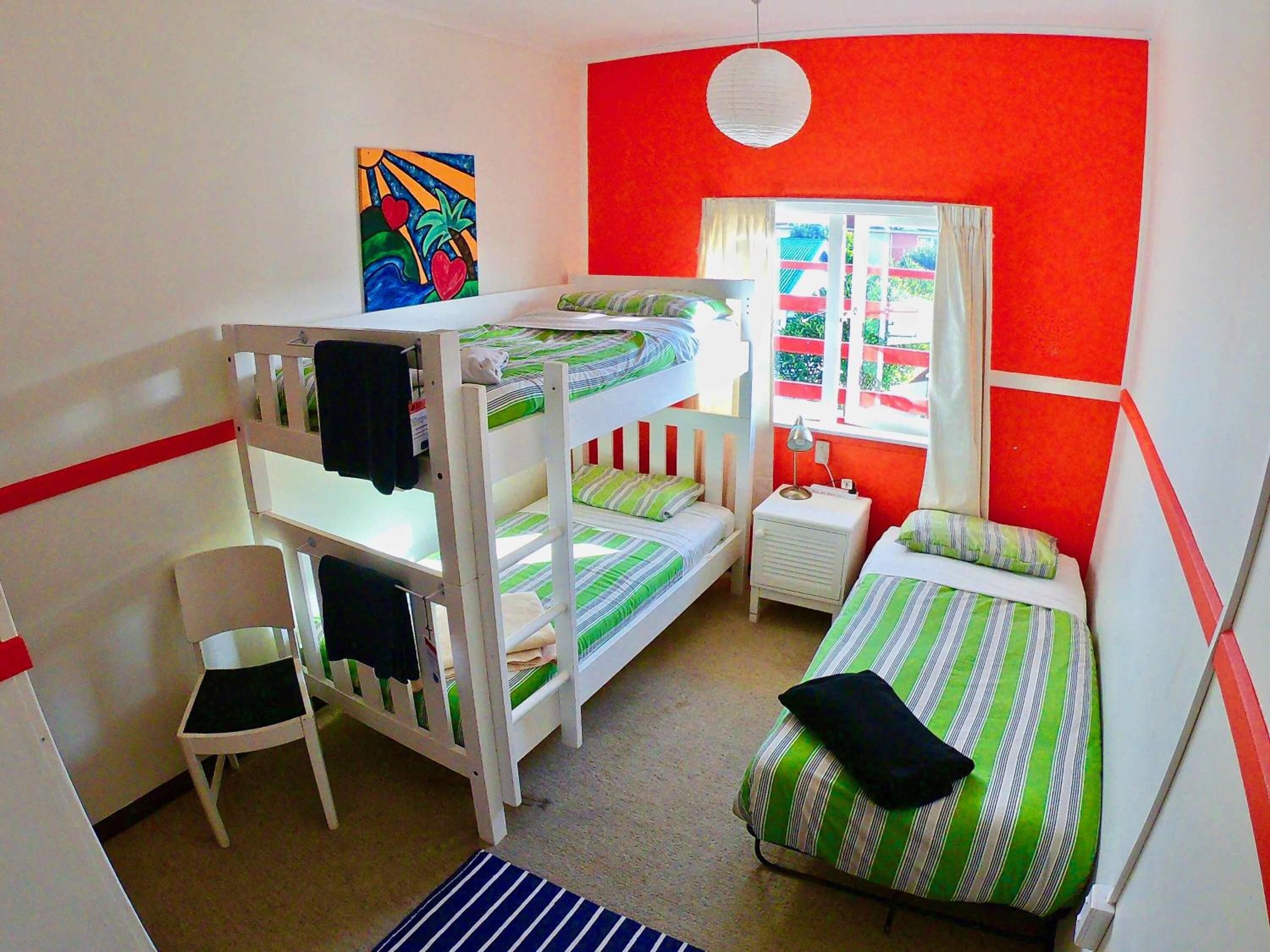 A dormitory room with bunk beds, a desk, a chair, and a window with curtains.