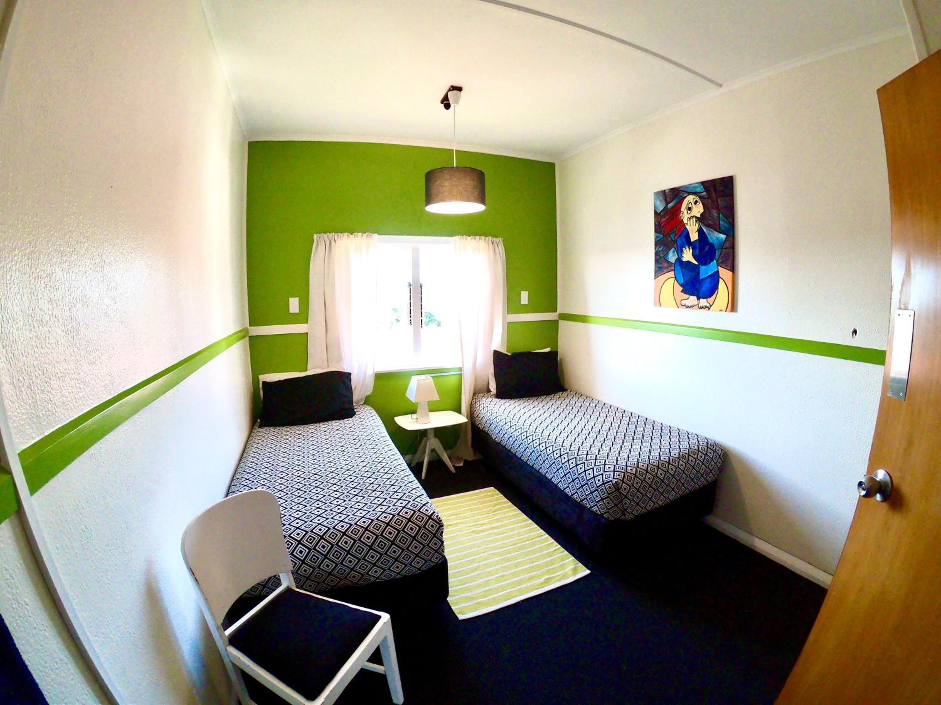 A small, single-room accommodation with two beds, a chair, a small table, and a lamp.