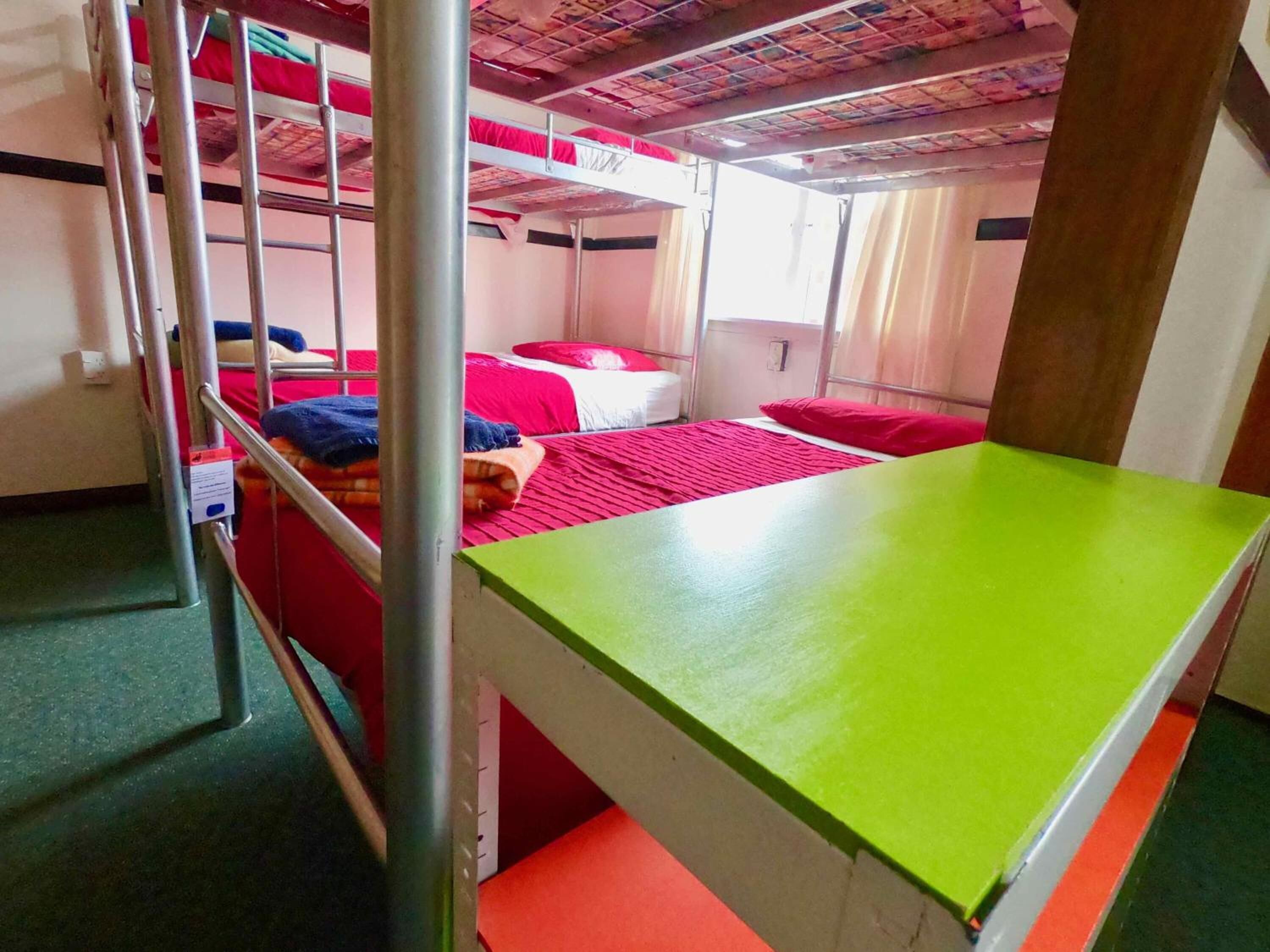 A dormitory room with bunk beds, a green table, and a red bedspread.