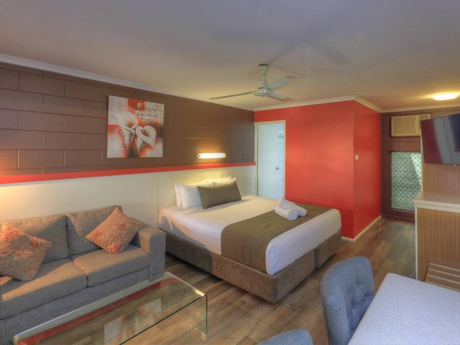 A compact hotel room with a bed, a sofa, a glass coffee table, a TV, and a ceiling fan.
