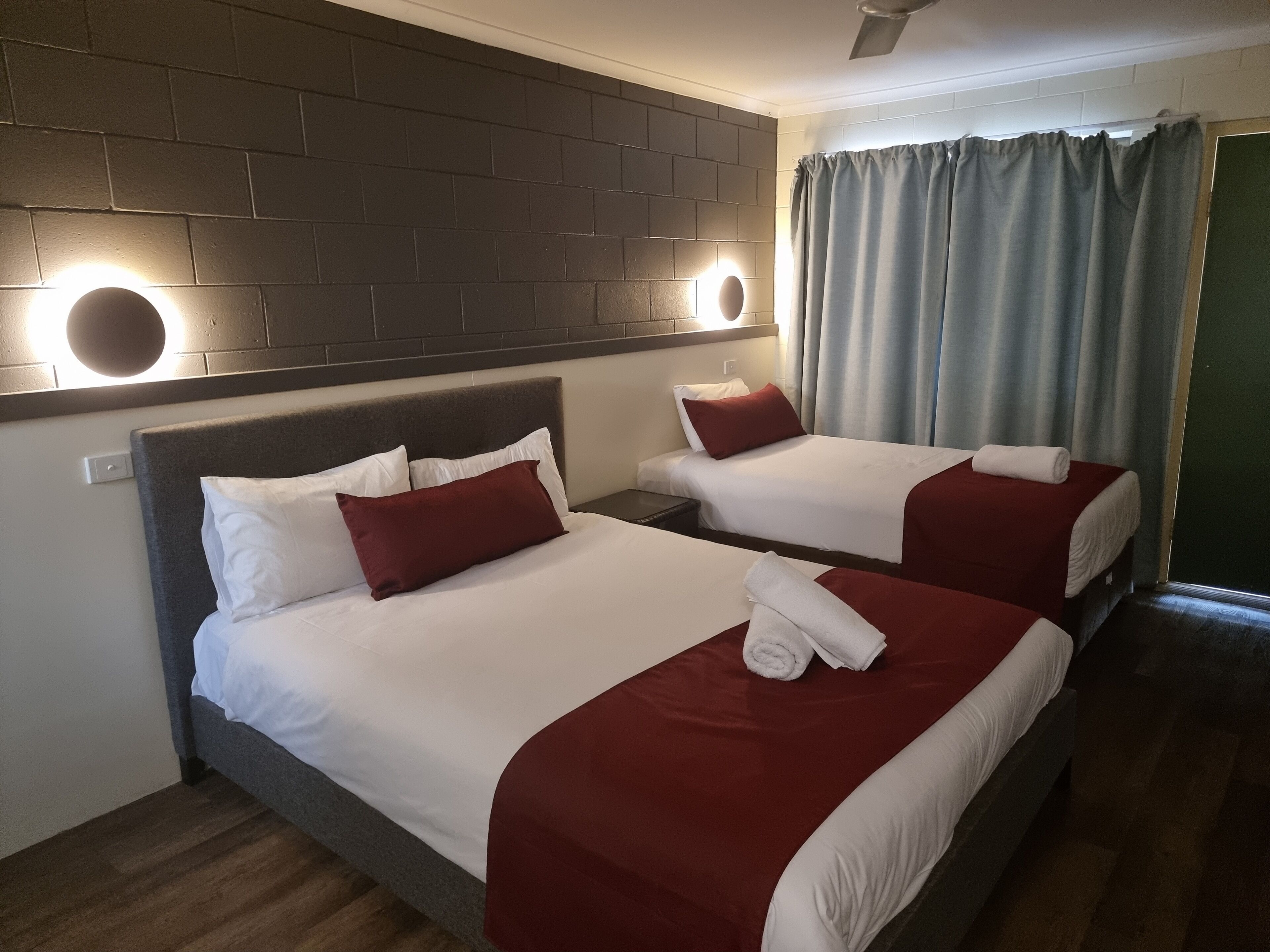 A hotel room with two beds, each with white and red bedding, a brick-patterned headboard, and a wall-mounted light fixture.
