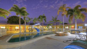A pool area with a slide, palm trees, and a building with a red roof. - Billabong Lodge Motel (Rosslea)