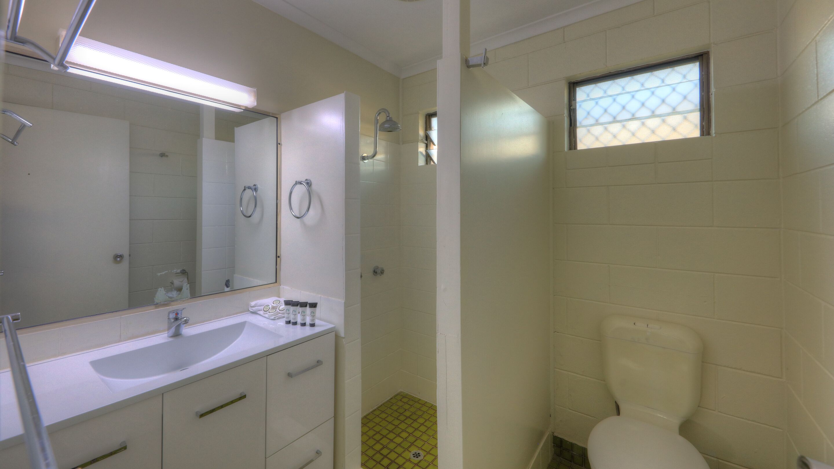 Un baño con lavamanos blanco, espejo e inodoro. Hay una zona de ducha con puerta de vidrio y una ventana sobre el inodoro.