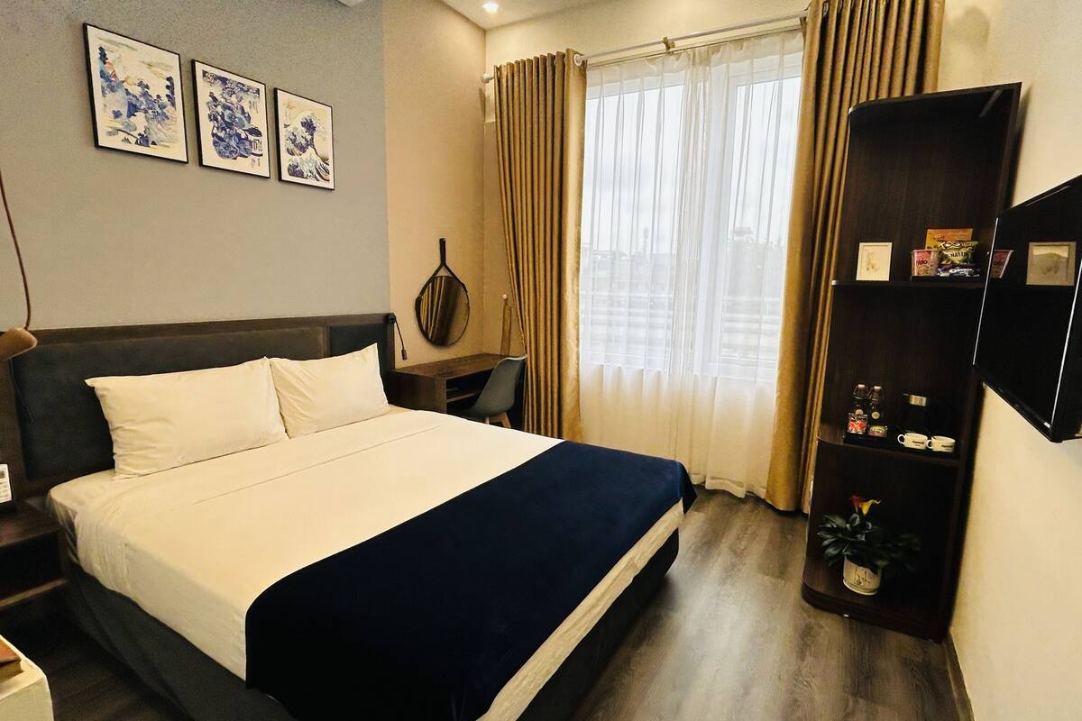 A hotel room with a bed, a desk, a chair, a TV mounted on the wall, and a shelf with various items.