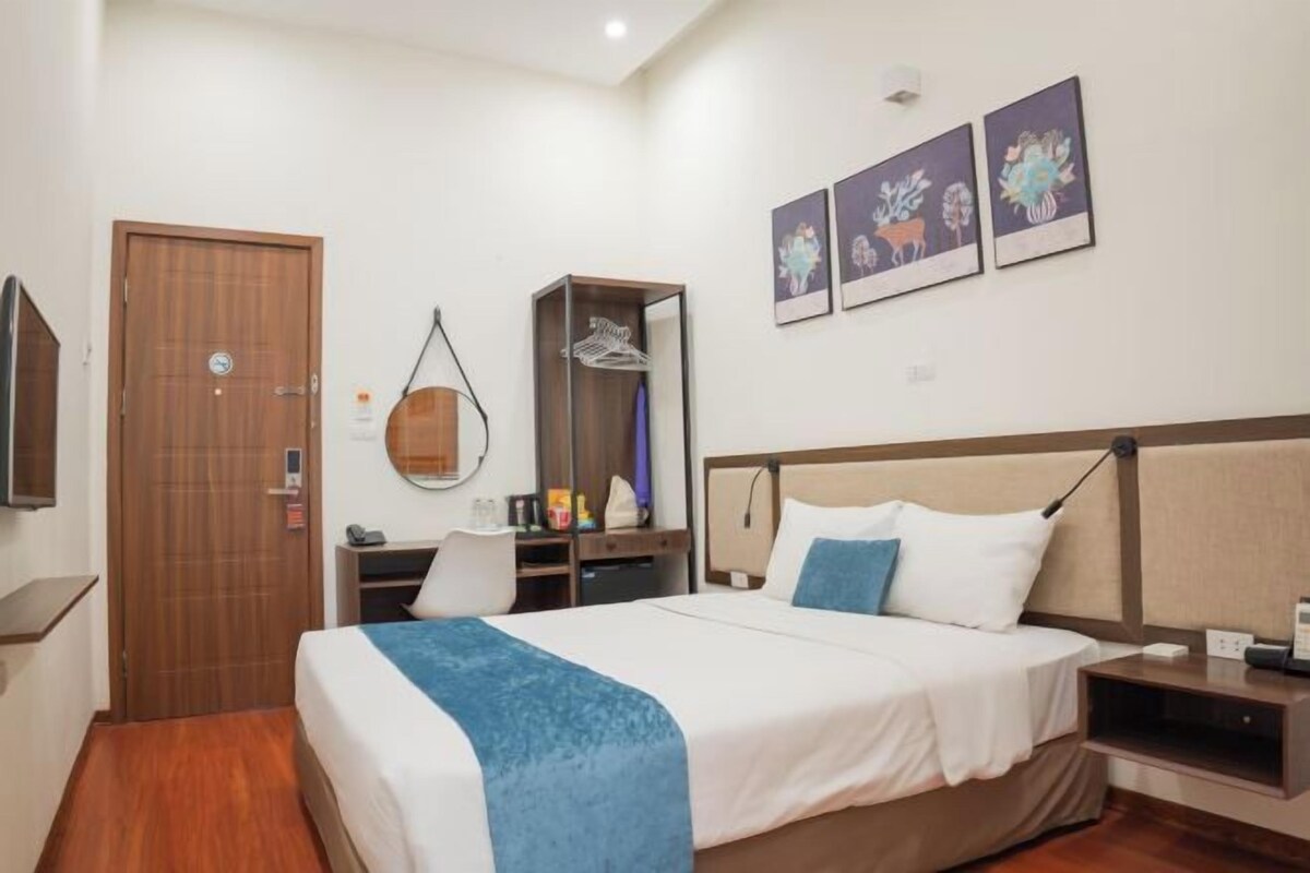 A hotel room with a bed, a desk, a chair, a wardrobe, and a mirror. There are three framed pictures on the wall.