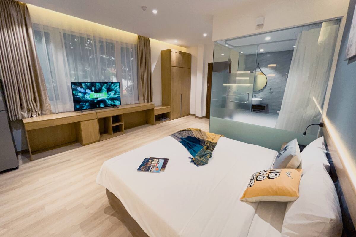 A modern hotel room with a large bed, a TV on a built-in shelf, a glass-enclosed bathroom, and a wardrobe.