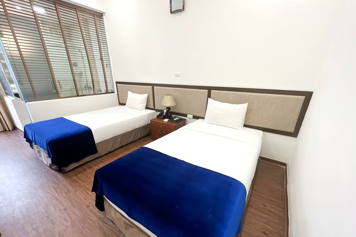 A hotel room with two beds, a nightstand, a lamp, and a window with blinds.