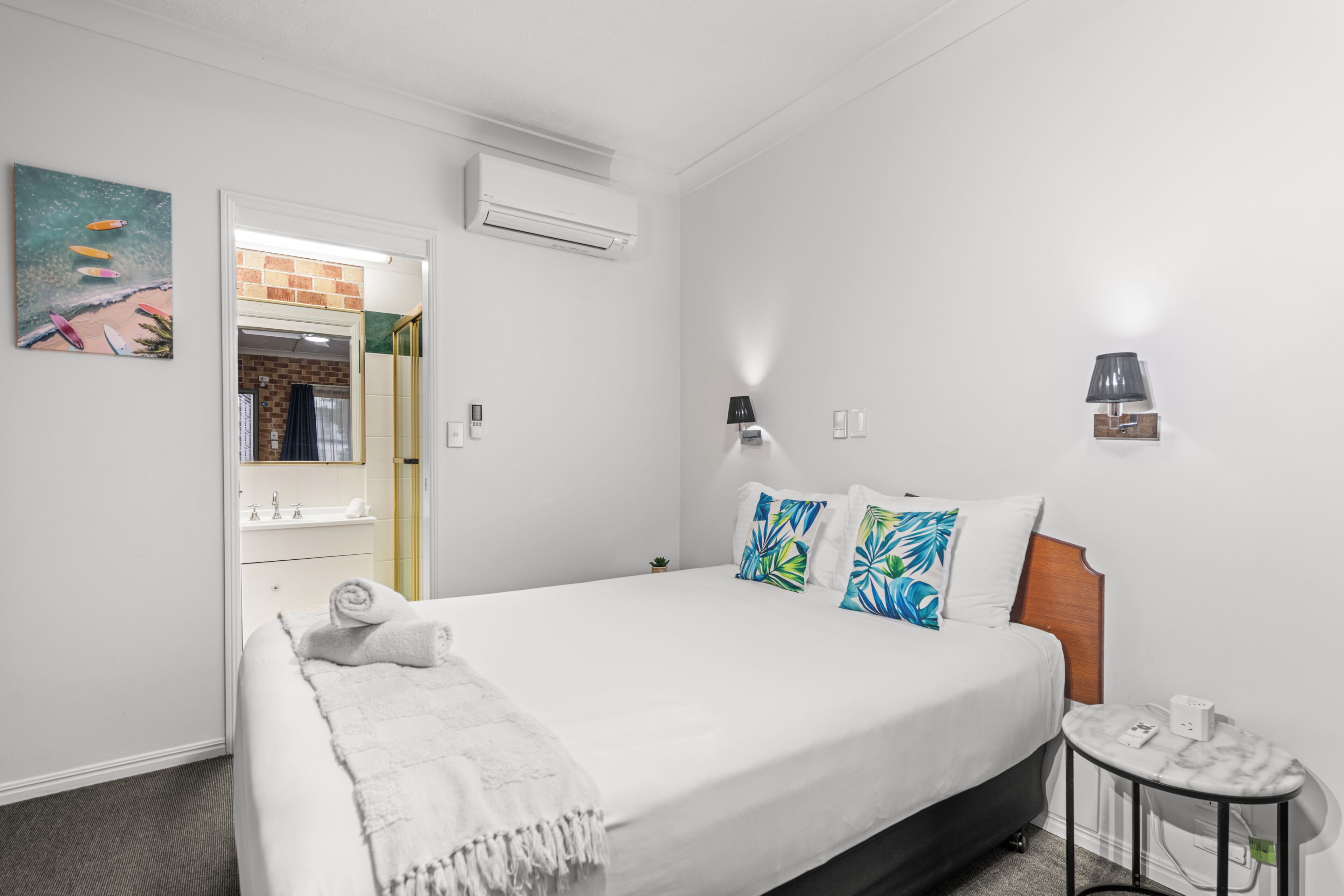 A hotel room with a bed, a small table, a wall-mounted lamp, and an air conditioning unit.
