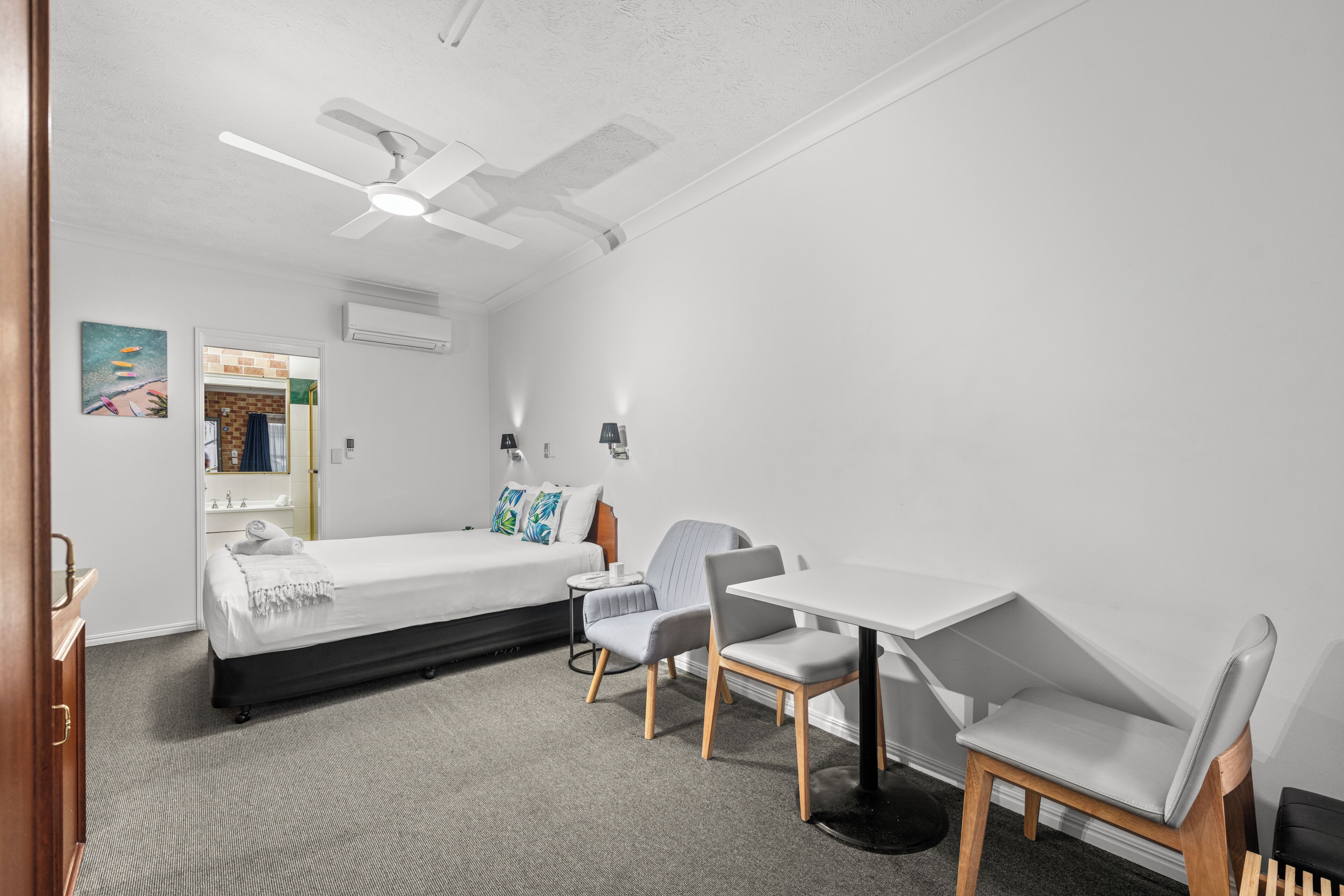 A hotel room with a bed, two chairs, a table, a ceiling fan, and a mirror.