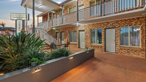 Un motel à deux étages avec une façade en briques, un balcon à l’étage supérieur et un espace extérieur aménagé avec des plantes, abrité par un auvent.