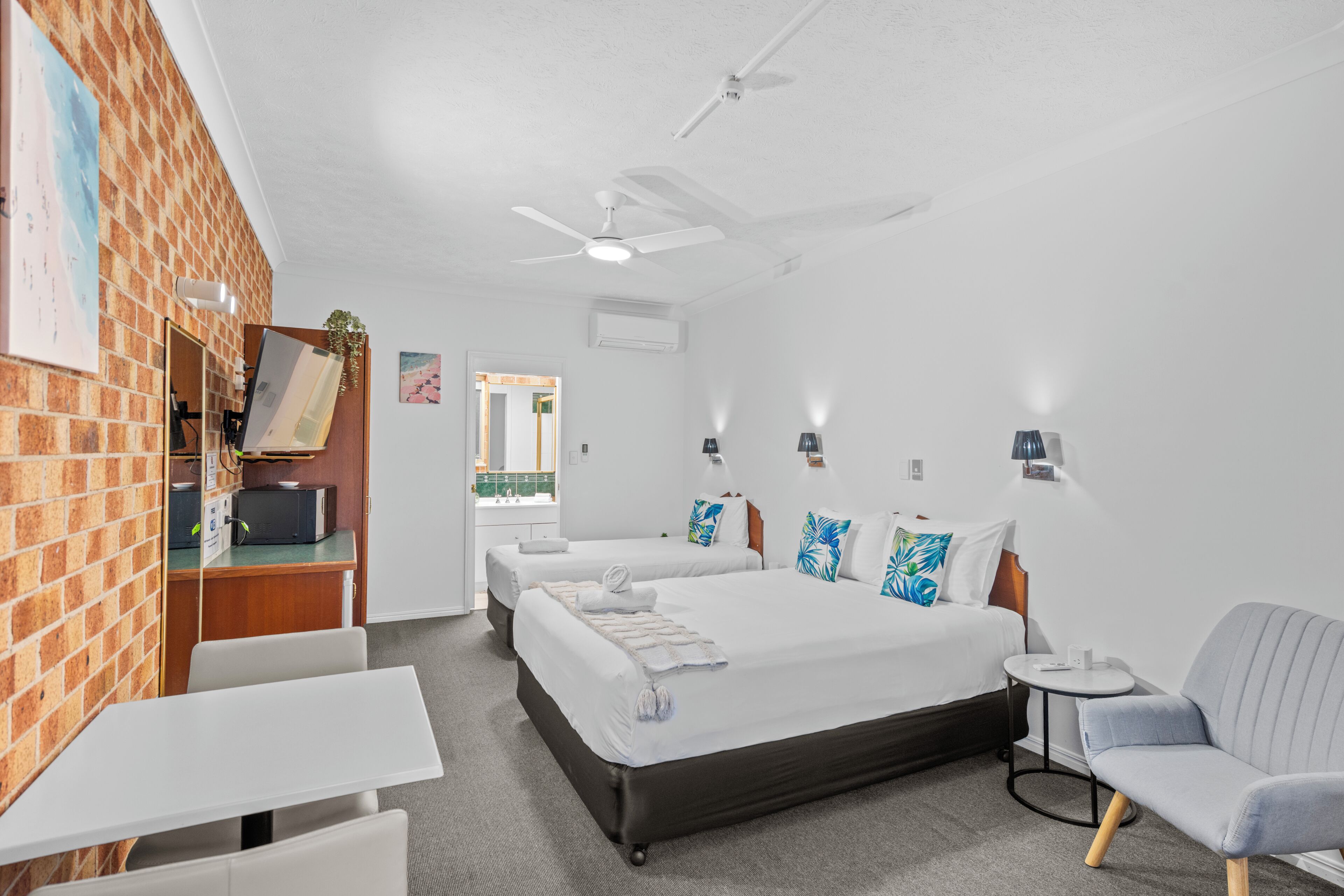A hotel room with a brick wall, two beds, a desk, a chair, and a ceiling fan.