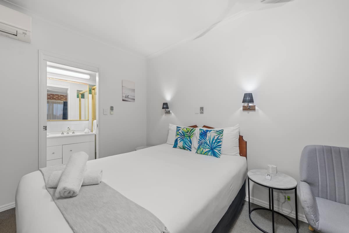 Een hotelkamer met een bed, een rond bijzettafeltje, een stoel, een spiegel, en een badkamer met een wastafel en spiegel.