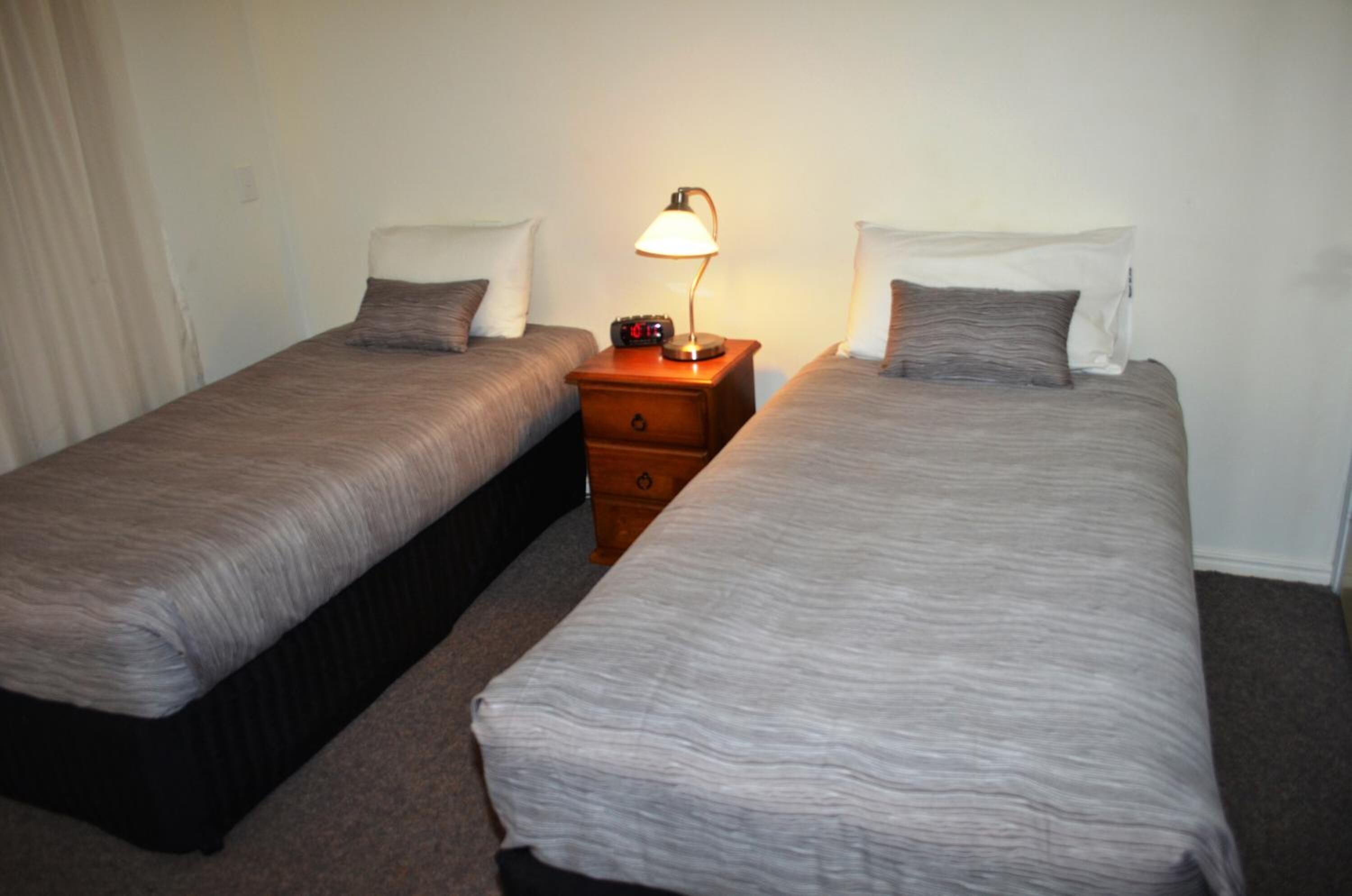 Two single beds with grey linens, a bedside table with a lamp, and a small clock.