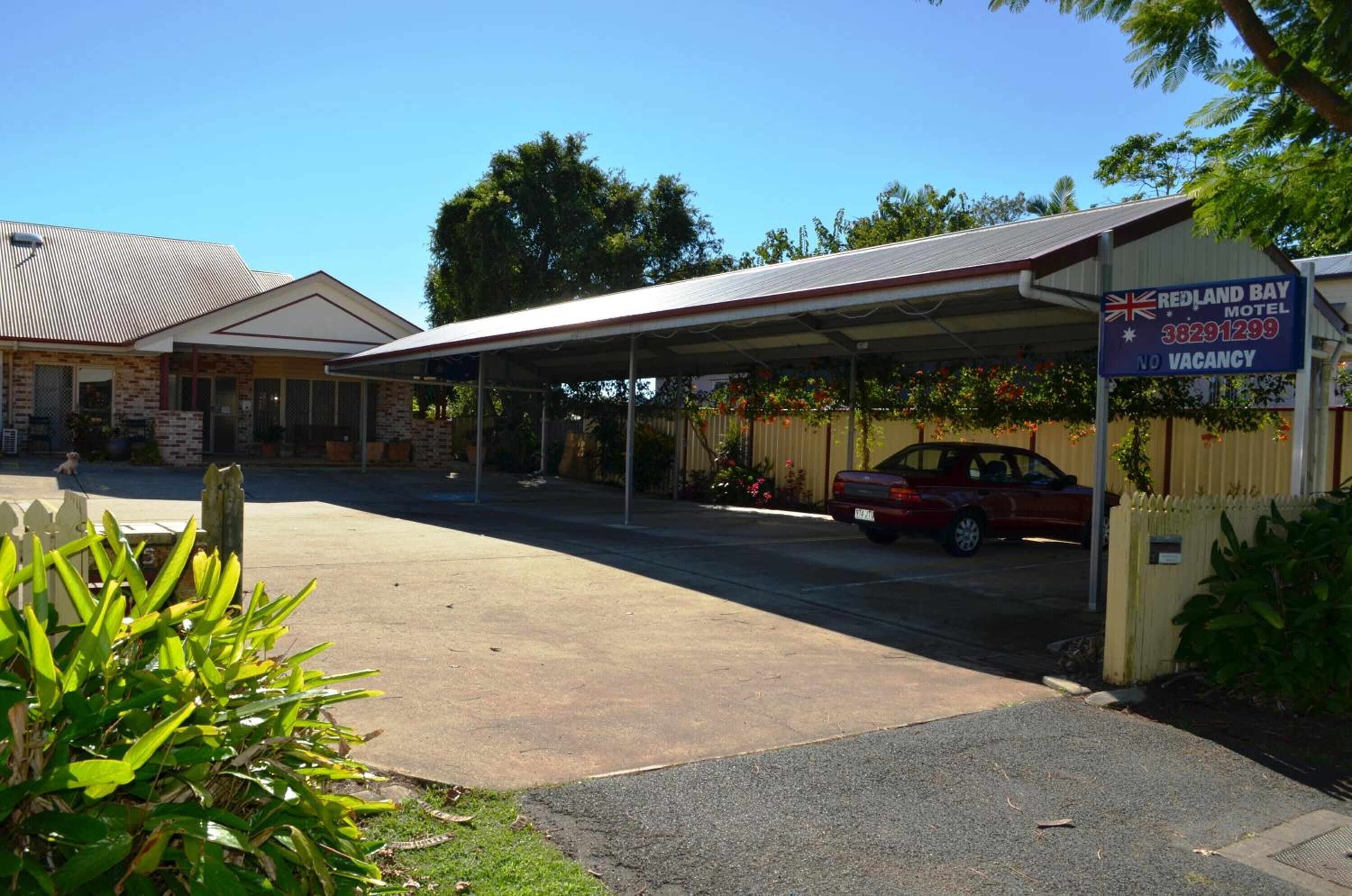 Un motel de Redland Bay doté d’un espace de stationnement couvert, avec une voiture rouge garée devant et un panneau indiquant qu’il n’y a plus de chambres disponibles.