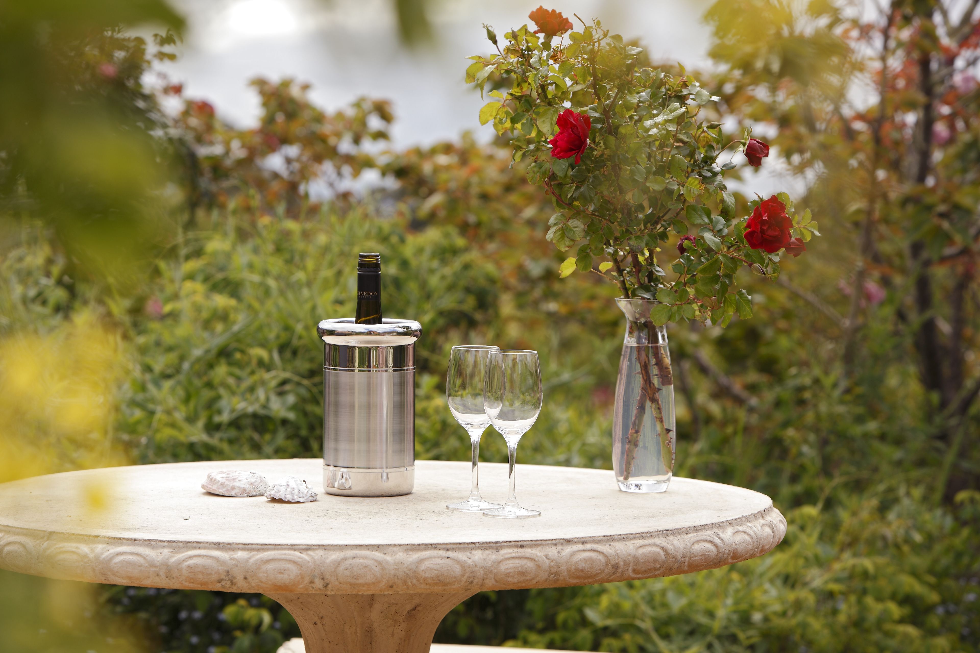 A round table with a metal container, two empty wine glasses, and a vase with red flowers.