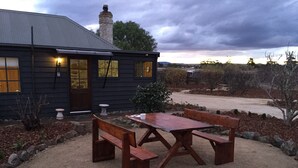 Et rustikt træhus med en grusbelagt terrasse, et træbord med bænke og en stensokkel.