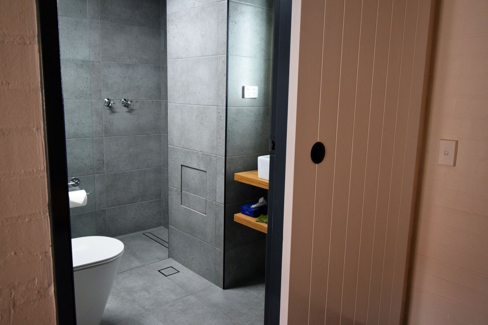 A modern bathroom with a toilet, shower, and a small shelf with toiletries.