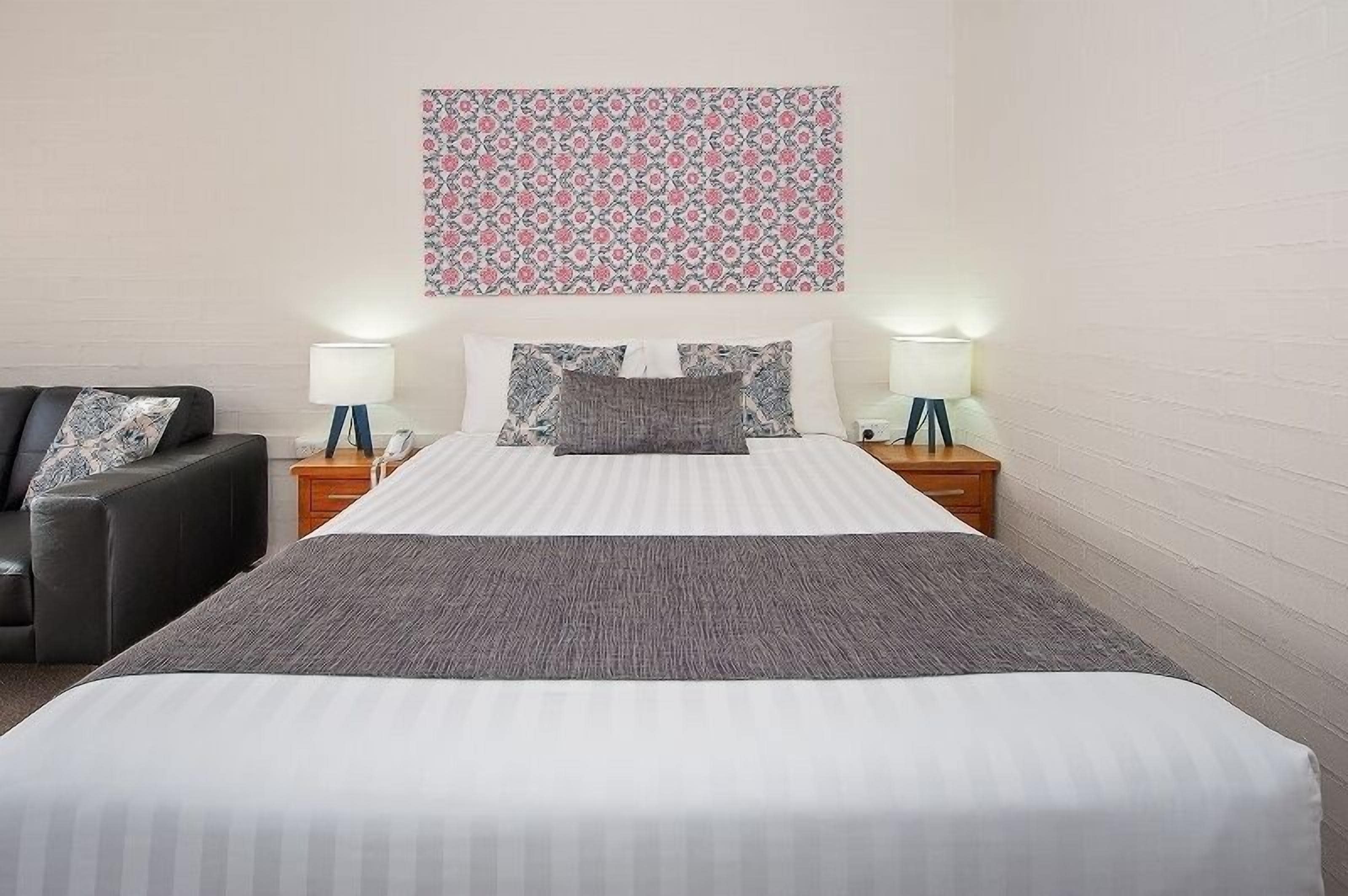 A neatly made bed with a striped white and gray duvet, two bedside lamps, a black leather sofa, and a floral-patterned wall hanging.